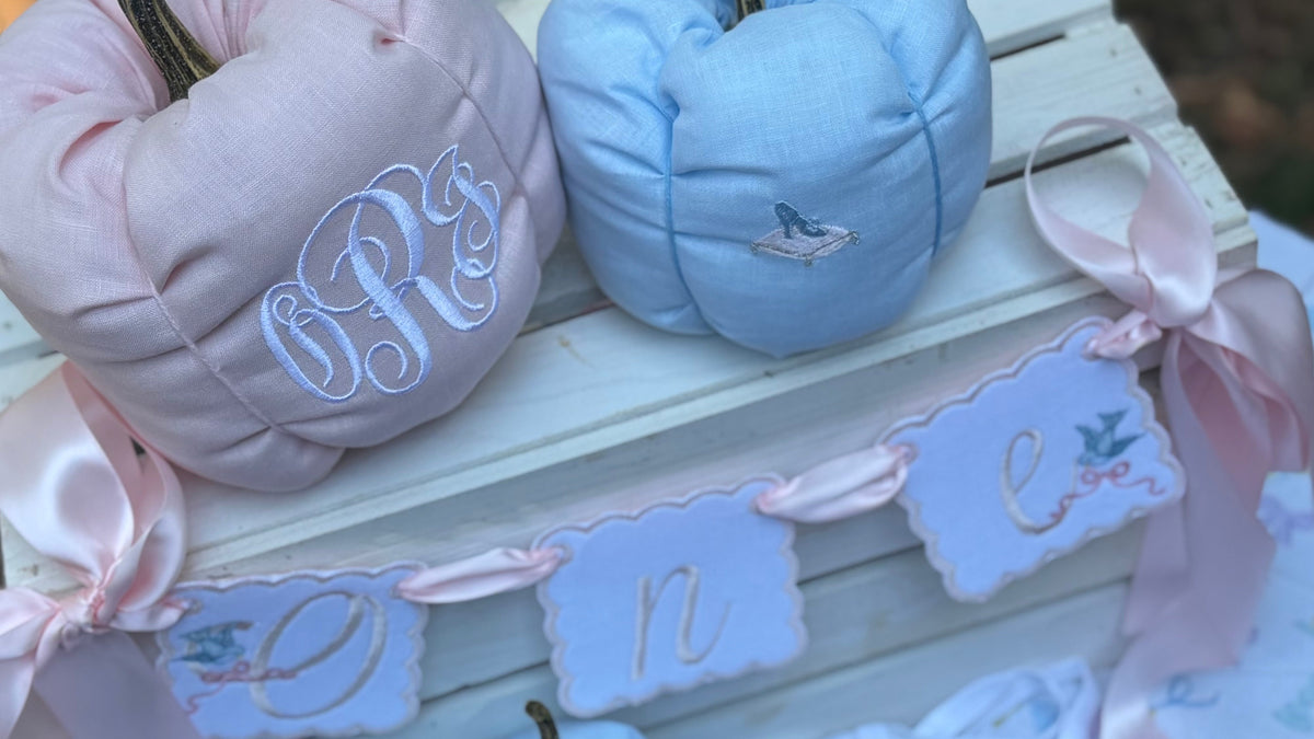 Personalized baby pumpkins in pastel pink and light blue, styled on a white wooden crate with a matching 'one' banner, pink satin ribbons, and a white bib with blue and pink floral details.