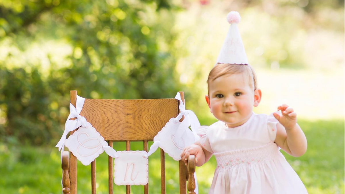 Child in a pale pink dress wearing a handmade white birthday party hat beside a chair with a personalized birthday banner